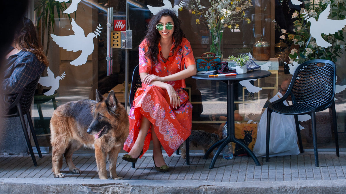 A woman in a vibrant red and orange patterned dress sits at an outdoor cafe table next to a German Shepherd dog, with white dove cutouts on a glass window in the background.